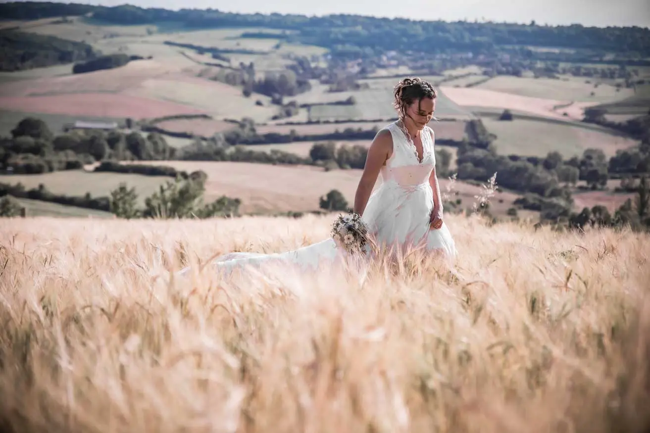 Mariée est dans un champs de blé avec son bouquet à la main.